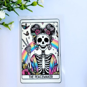 May include: A colorful sticker featuring a skeleton with a floral crown, giving a peace sign. A rainbow and butterflies are in the background. The text "THE PEACEMAKER" is at the bottom of the sticker.