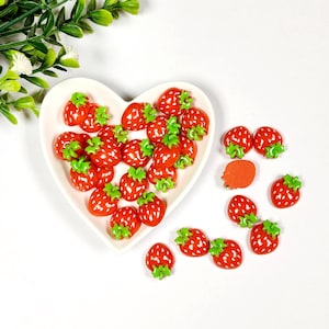 May include: A collection of red strawberry-shaped embellishments with green leafy tops. Some are arranged in a heart-shaped dish, while others are scattered on a white surface. The strawberries have white speckles.