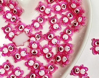May include: A collection of small, pink, bear-shaped resin charms with white bellies and black eyes, arranged in a heart-shaped dish. The charms have a glittery finish, and some are paired together.