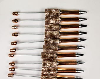 May include: An overhead shot displays ten pens. Each pen has a silver shaft, a gold-colored tip, and a gold-colored barrel. The barrel is adorned with small, sparkling, gold-colored rhinestones. The pens are arranged on a white surface.