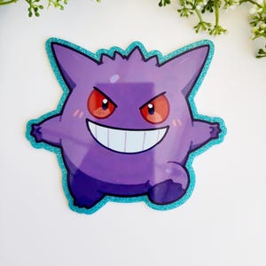 May include: A purple Gengar character sticker with a wide, toothy grin and glowing red eyes. The sticker features a distinct, shimmering teal glitter border that outlines the creature's spiky, rounded shape against a plain white background.