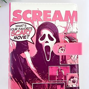 May include: A rectangular notebook featuring a pink and white graphic print inspired by the movie Scream. The cover displays the text SCREAM, a speech bubble with the text WHAT'S YOUR FAVORITE SCARY MOVIE?, and several comic-style panels.