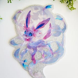 May include: A die-cut sticker featuring a stylized pink and purple creature with long ears and flowing ribbons, set against a large, pale circular moon background. The sticker has a distinct silver glitter border and a shimmering, holographic finish.
