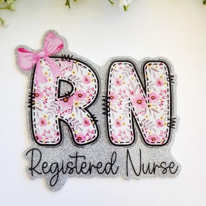 May include: A silver glitter cutout featuring the letters RN with a pink floral pattern and a pink bow on the R. Below the letters, the text Registered Nurse is written in black script. The design has a stitched border effect throughout.