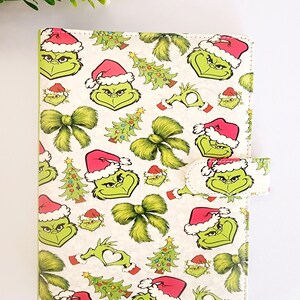 May include: A rectangular notebook cover featuring a repeating pattern of green Grinch faces wearing red Santa hats, decorated Christmas trees, and fuzzy green bows on a white background, with a matching magnetic closure tab on the right side.