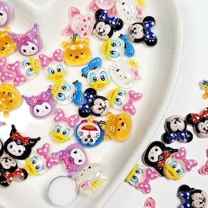 May include: A collection of colorful resin charms in various cartoon character designs, including Mickey Mouse, Minnie Mouse, Donald Duck, and Hello Kitty. The charms are arranged in a white heart-shaped dish, with a glossy finish.