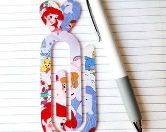 May include: A decorative paperclip featuring Disney princess characters, including Ariel and Cinderella, with a white and gray pen resting on lined paper. The paperclip has a heart-shaped top and is adorned with various cartoon images.