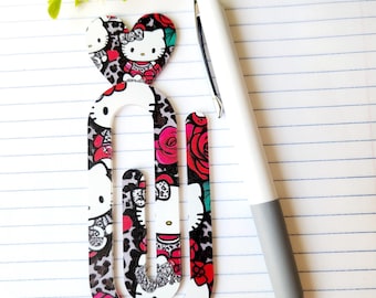 May include: A decorative paperclip featuring a heart-shaped top and a U-shaped design. The paperclip showcases a pattern of Hello Kitty, roses, and leopard print. A white and gray pen rests beside the paperclip. The text "My Lovely Creations" is at the bottom.