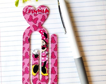 May include: A pink Minnie Mouse paperclip with a heart-shaped top and bow pattern. The paperclip features a Minnie Mouse illustration and the word "MINNIE". A white and gray pen rests on the lined paper.