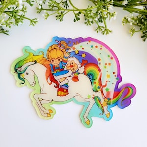 May include: A die-cut acrylic sticker featuring a cartoon character with blonde hair and a small white creature riding a white unicorn with a rainbow mane and tail. The design includes a colorful rainbow arc and scattered yellow and orange stars.