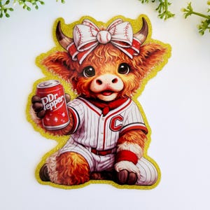 May include: A die-cut sticker featuring a fluffy brown Highland cow wearing a white baseball uniform with red pinstripes and a letter C on the chest. The cow has a baseball-themed bow on its head and holds a red soda can labeled Dr Pepper.