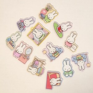 May include: A collection of twelve flat acrylic charms featuring the white rabbit character Miffy in various scenes, including reading, dressing, and sitting in a room, each with a small hole for hanging and pastel-colored graphic illustrations.