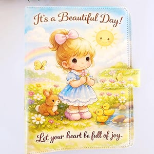 May include: A rectangular notebook cover featuring a cartoon illustration of a child with blonde hair, a blue dress, and a pink bow. The scene includes a rabbit, two yellow ducks, a sun, and a rainbow. Text reads: It's a Beautiful Day! Let your heart be full of joy.
