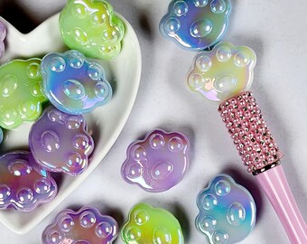 May include: Iridescent paw-shaped beads in shades of green, purple, and blue are scattered on a white heart-shaped dish and a light gray surface. A pink pen with a bedazzled handle is in the background. The beads have a glossy finish.