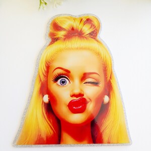 May include: A die-cut sticker featuring a stylized illustration of a person with bright yellow hair styled in a high bun. The face has one wide blue eye, one winking eye, and bold red lips. The sticker has a silver glitter border and a glossy finish.
