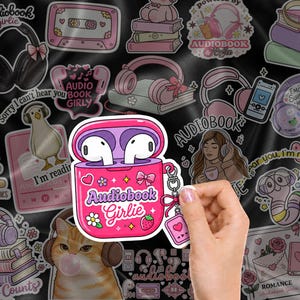 May include: A pink sticker featuring a charging case for wireless earbuds with white earbuds inside. The case is decorated with a bow, a strawberry, a flower, and the text Audiobook Girlie. A small charm with a heart icon hangs from the side.