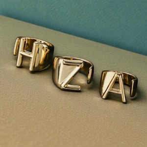 以下が含まれることがあります： H、Z、Aの文字がそれぞれデザインされた3つのゴールドカラーのリング。大胆なブロックレターのデザインと、磨かれた仕上がりが特徴です。ニュートラルな表面に、ティールブルーの背景で展示されています。