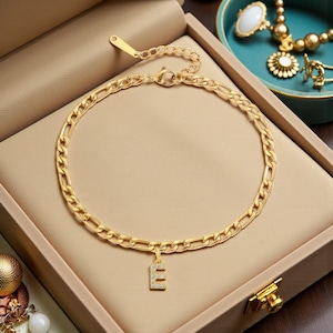 Könnte beinhalten: Ein goldfarbenes Fußkettchen mit Kettenglied-Design. Das Fußkettchen hat einen kleinen Anhänger in Form des Buchstabens "E", der mit kleinen, klaren Edelsteinen besetzt ist. Das Fußkettchen wird in einer beigen Schmuckschatulle präsentiert.