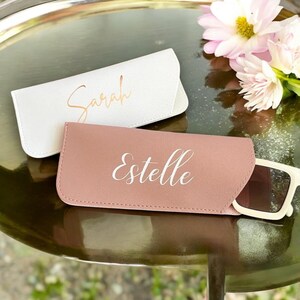 May include: Two personalized eyeglass cases, one white with the name "Sarah" and one pink with the name "Estelle" in white lettering.
