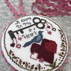 May include: A round, cross-stitched embroidery piece with the text "A Song In My Heart" and a key design. The design includes a blue bird, a heart, and floral accents. It has a pink scalloped edge and a pink ribbon for hanging.