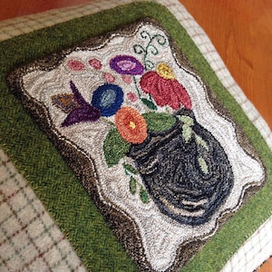 May include: A green and brown wool pillow with a white frame featuring a needlepoint design of a black vase with colorful flowers. The pillow has the text "Three Sheep Studio.com" at the bottom.