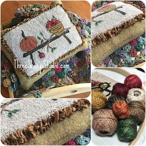 May include: A hand-stitched wool pillow with a fall-themed design featuring a pumpkin, a bird in a nest, and a branch. The pillow is trimmed with brown ruffled fabric and is displayed on a wooden tray with a background of colorful fabric rosettes. The image also shows a close-up of the pillow's edge and a collection of yarn balls in various fall colors.