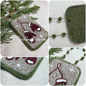 May include: A hand-stitched wool ornament featuring a pair of red mittens with white trim, hanging from a green wool loop. The ornament is decorated with white snowflakes on a beige background and has a green wool border. The ornament is attached to a green wool chain with brown pom-poms.
