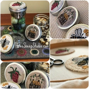 May include: A collection of needlepoint embroidery hoops with various designs, including a strawberry, a house, and a bee. The hoops are displayed on a table with a variety of buttons and other craft supplies.