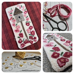 May include: A close-up of a needlepoint project depicting a birdhouse with pink flowers. The project is on a white background and is surrounded by a variety of embroidery supplies, including yarn, scissors, and a needlepoint canvas.
