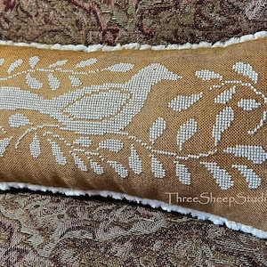 May include: A brown linen pillow with a white embroidered design of a bird and leaves. The pillow has a white fringe border. The text "Three Sheep Studio.com" is embroidered on the bottom right corner.