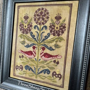 May include: Framed cross-stitch artwork featuring a central floral design with two red birds. The piece includes burgundy, blue, and green accents on a beige background. The black frame has a beaded edge and the text "ThreeSheepStudio.com".