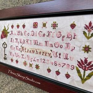 May include: A cross-stitch sampler with a white background and pink and green embroidery. The sampler features the alphabet, numbers, and a strawberry design. The text "Three Sheep Studio.com" is at the bottom of the sampler.
