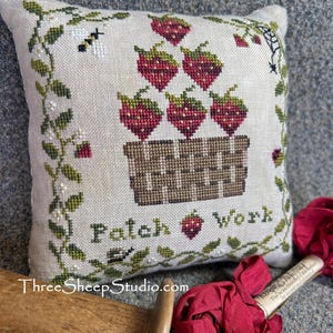 May include: A cross-stitched pillow with a basket of strawberries. The pillow has a cream background with green vines and a bee. The words "Patch Work" are stitched below the basket. The website ThreeSheepStudio.com is at the bottom.