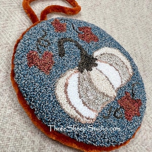 May include: A round punch needle wall hanging featuring a white pumpkin with a dark brown stem on a textured blue background. The design includes small red-orange leaves and the text ThreeSheepStudio.com. It has a fuzzy orange border.