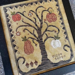 May include: Framed cross-stitch sampler featuring a tree with red apples, a pear, and a white apple. The sampler has a black border and is framed in a black wooden frame. The text "ThreeSheepStudio.com" is visible.
