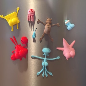 May include: A collection of colorful cartoon character magnets, including a yellow sponge, a pink jellyfish, a brown bear-like figure, a red crab, a blue squid, a pink starfish, and a blue object. The magnets are arranged on a metallic surface.