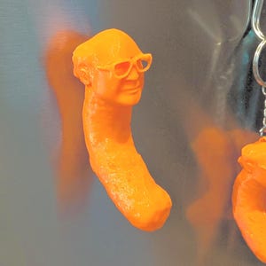 May include: Orange novelty keychain featuring a caricature head wearing glasses. The head is attached to a long, curved body. The keychain is made of a solid, smooth material and is set against a silver background.