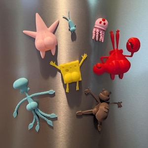 May include: A collection of colorful cartoon character magnets, including a pink starfish, a yellow sponge, a red crab, a blue squid, a pink jellyfish, and a small blue fish. The magnets are arranged on a metallic surface.