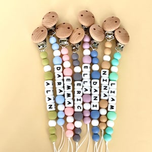 May include: A collection of personalized baby pacifier clips with wooden and metal clasps. Each clip features a string of colorful silicone beads and white lettered blocks spelling out names like "Alan", "Dara", and "Emma".