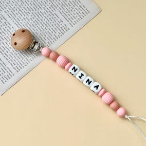 以下が含まれることがあります： ピンクと白のシリコンビーズに、黒い文字で「NINA」と綴られています。ビーズは木製クリップとシリコン製の歯固めにつながっています。