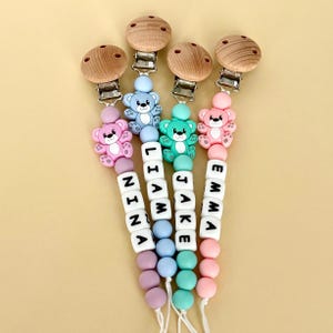 Puede incluir: Cuatro clips para chupete de bebé con cierres de madera y metal. Cada clip presenta un dije de oso colorido y una cadena de cuentas que deletrean un nombre: Nina, Liam, Jake y Emma. Los clips son en tonos rosa, azul y verde.