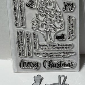May include: Clear stamp set with Christmas-themed sentiments, including "Merry Christmas," "Peace," and "Joy." Features a heart-shaped tree design and a dog illustration. Includes a metal die cut of a Christmas tree.