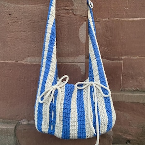 May include: A blue and cream striped crochet shoulder bag with long straps. The bag has a rounded shape and is tied with cream-colored yarn at the top. The bag is made of a thick yarn and has a textured appearance.
