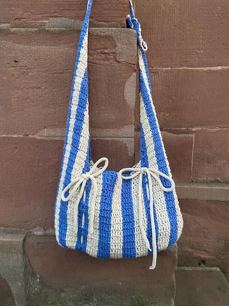 May include: A blue and cream striped crochet shoulder bag with long straps. The bag has a rounded shape and is tied with cream-colored yarn at the top. The bag is made of a thick yarn and has a textured appearance.
