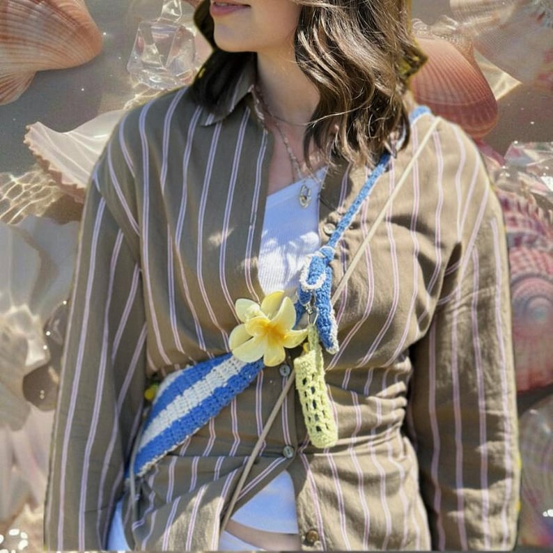 May include: A handmade crochet crossbody bag with a blue and white striped design, adorned with a yellow flower. The bag has a long strap and a small, light green crochet pouch. The background features a striped shirt.