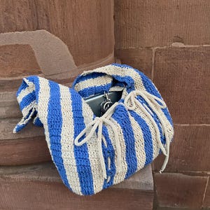 May include: A blue and beige striped crochet bag with a drawstring closure. The bag is open, revealing a phone and keys inside. The bag is resting on a stone surface.