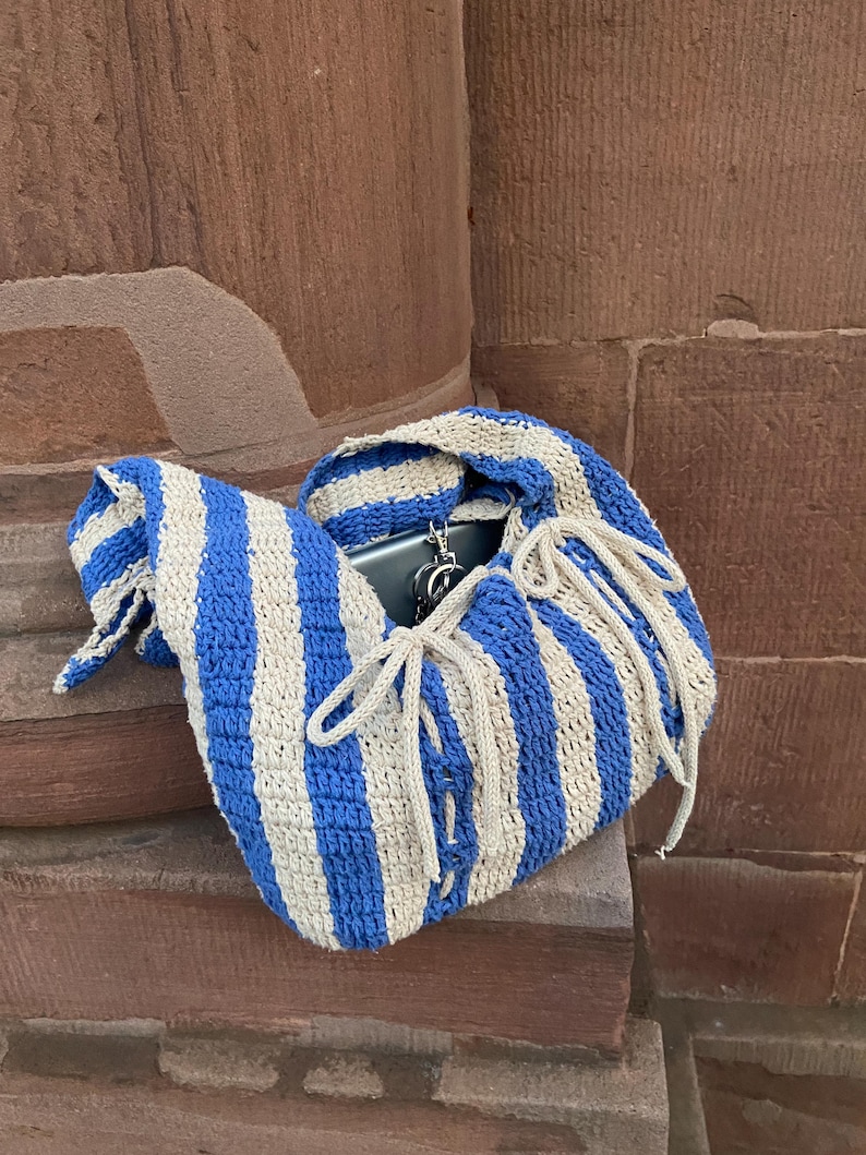 May include: A blue and beige striped crochet bag with a drawstring closure. The bag is open, revealing a phone and keys inside. The bag is resting on a stone surface.