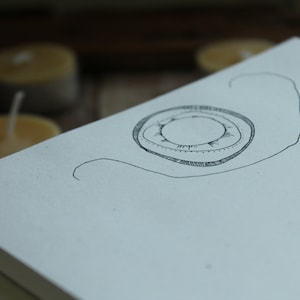 May include: A white notebook with a black ink drawing of a circle with a smaller circle inside. The drawing is on the top of the notebook, which is stacked on top of other notebooks.