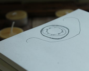 May include: A white notebook with a black ink drawing of a circle with a smaller circle inside. The drawing is on the top of the notebook, which is stacked on top of other notebooks.