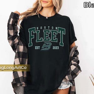 May include: Black t-shirt with the text "BOSTON FLEET" in green and white lettering. The shirt also features the text "EST 2023" and a stylized "B" logo. The shirt is worn with a plaid shirt.